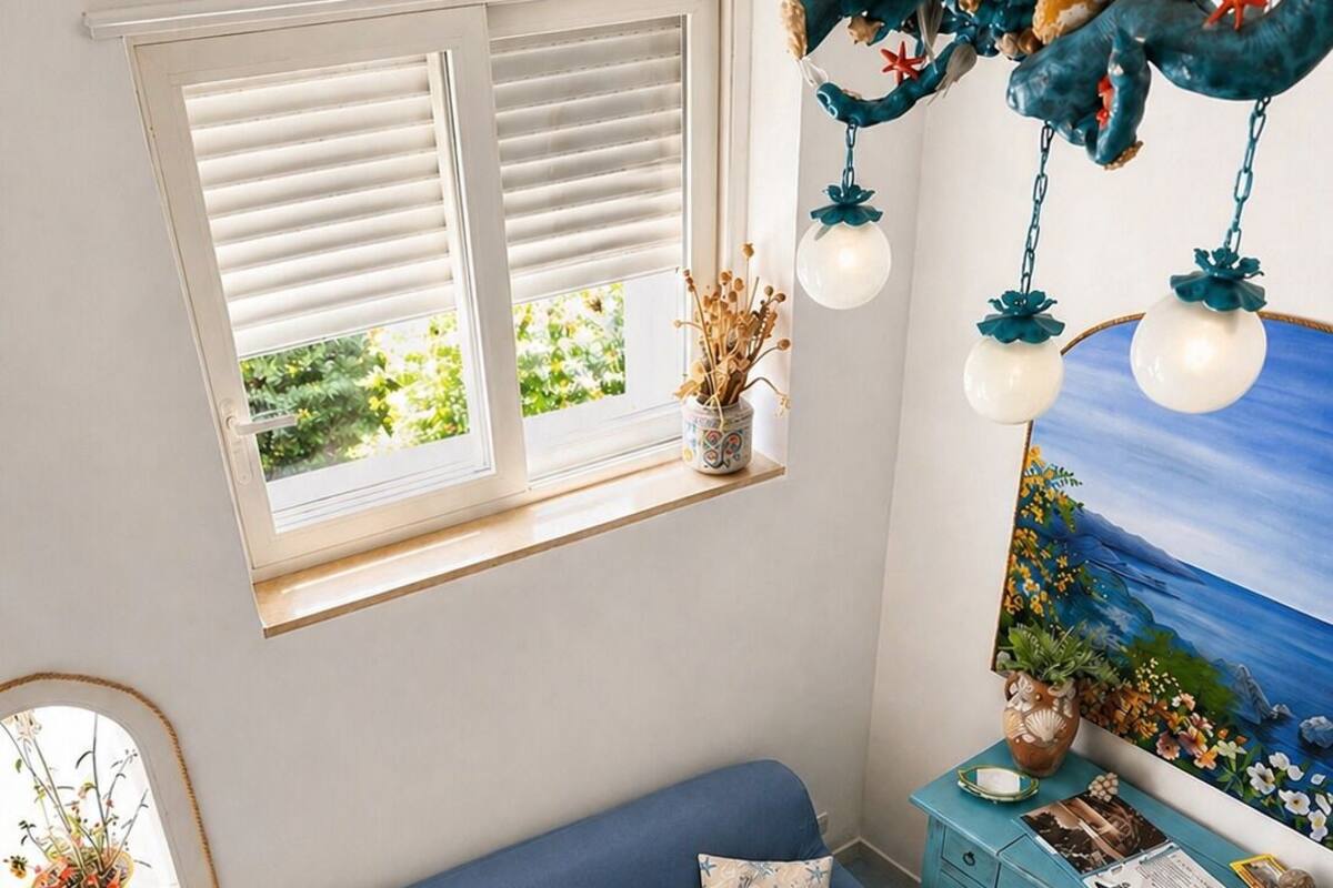 Une pièce avec un canapé bleu, une petite table en bois, une commode, une fenêtre avec des stores et un élément de décoration suspendu.