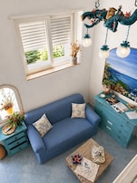 Ein Zimmer mit einem blauen Sofa, einem kleinen Holztisch, einem Schminktisch, einem Fenster mit Jalousien und einer Wanddekoration.