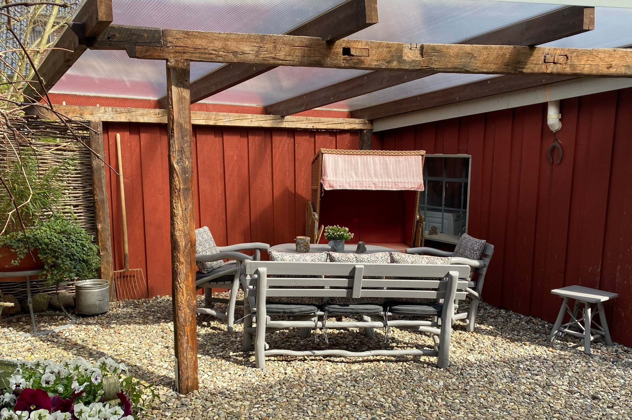 A covered outdoor seating area with a wooden bench, a table, and a window with a pink awning.
