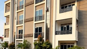 A multi-story residential building with balconies, a paved driveway, and landscaped areas with small trees.