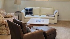Een woonkamer met een beige bank, een bruine fauteuil en een kleine tafel. Er zijn twee ramen met jaloezieën, en een lamp is zichtbaar.