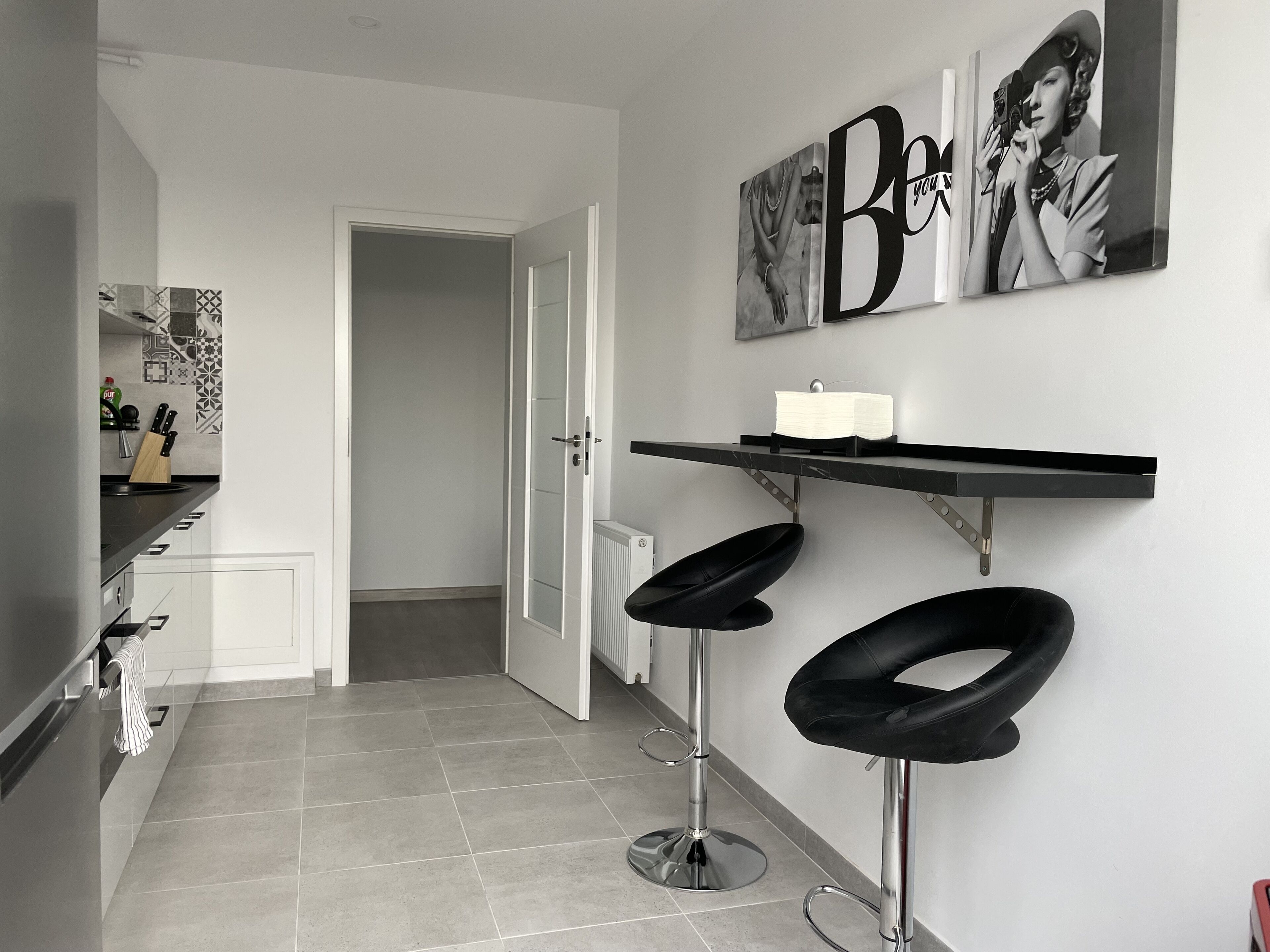 Eine moderne Küche mit weißen Schränken, einer schwarzen Arbeitsplatte und einer gefliesten Spritzschutzwand. Zwei schwarze Barhocker mit Metallbeinen stehen bereit. Eine weiße Tür führt in einen weiteren Raum.