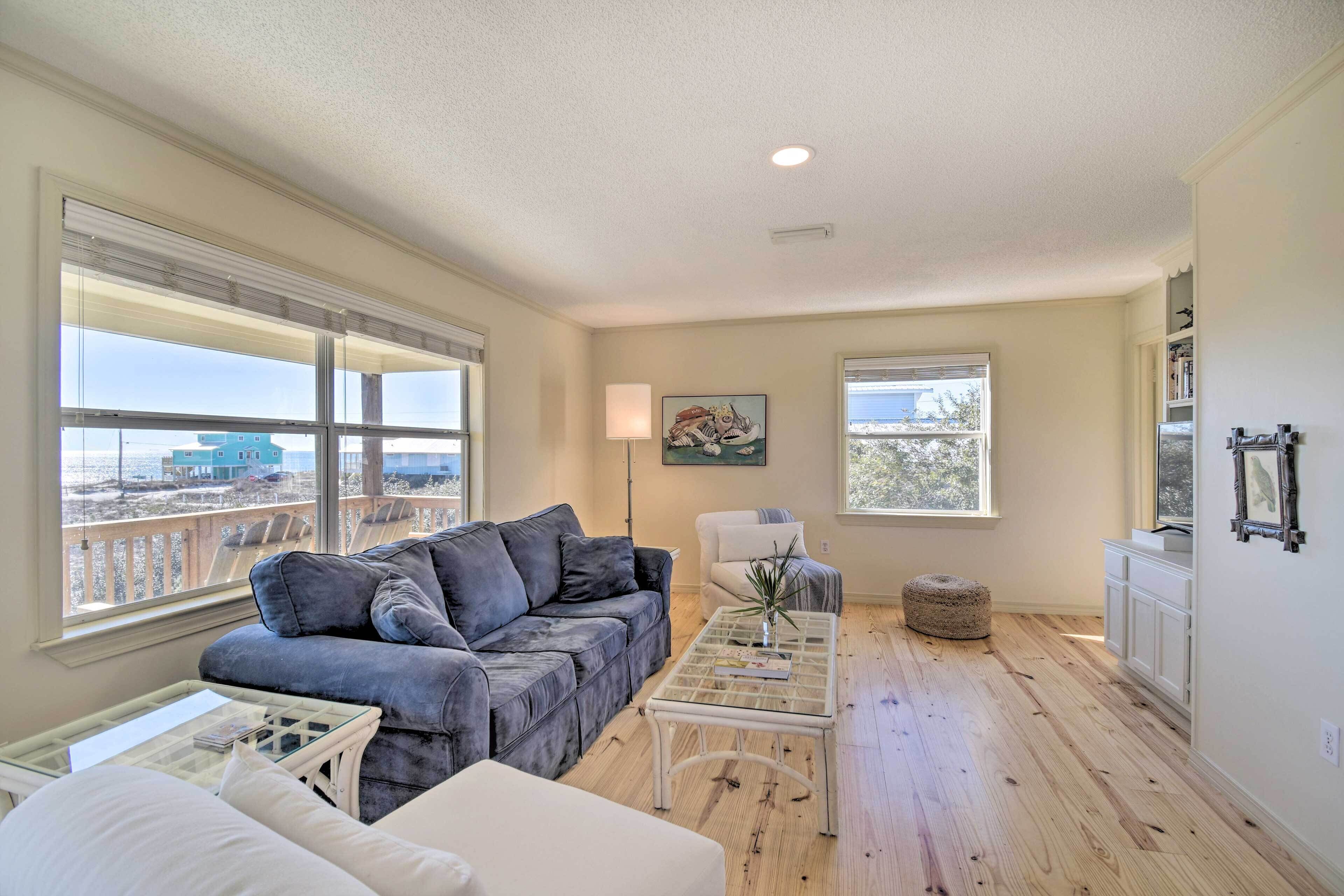 A spacious living room with a grey sofa, a white armchair, a glass coffee table, and a view of the beach through large windows.