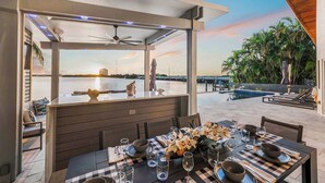 An outdoor dining area with a view of the water and sunset, a covered seating area, and a poolside lounge. - Bayfront Escape Poolside Luxury Villa with Sunset Views (North Bay Village)