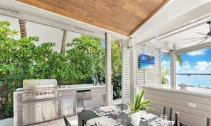 An outdoor kitchen with a grill, dining table set for four, and a view of the sea.