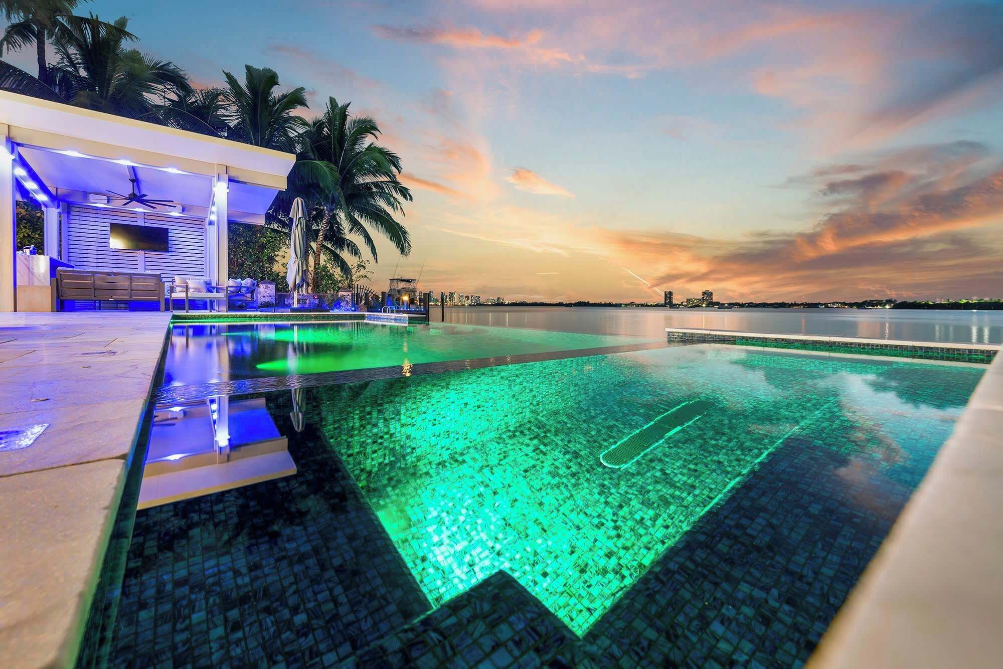 Une piscine offrant une vue sur le coucher de soleil, des palmiers et un bâtiment doté d’un espace aménagé au bord de la piscine.