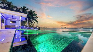 A swimming pool with a view of the sunset, palm trees, and a building with a poolside area. - Bayfront Escape Poolside Luxury Villa with Sunset Views (North Bay Village)