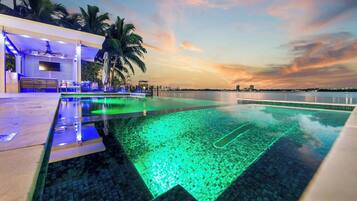 Une piscine offrant une vue sur le coucher de soleil, des palmiers et un bâtiment doté d’un espace aménagé au bord de la piscine.