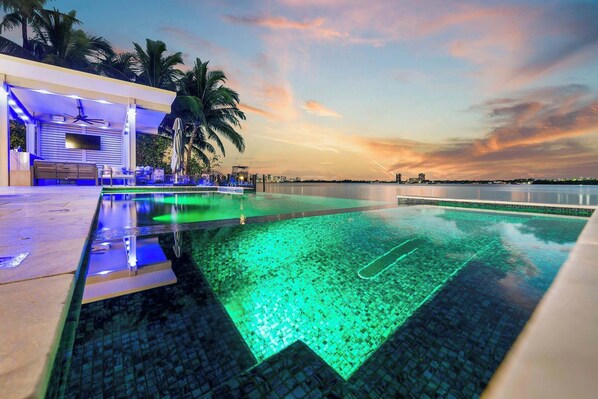 A swimming pool with a view of the sunset, palm trees, and a building with a poolside area.