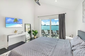 A bedroom with a bed, a desk, a TV, a plant, and a view of the sea.