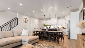 A modern kitchen with white cabinets, a dining area with a wooden table and chairs, and a beige sectional sofa with a patterned rug. - Desert Daydream: Desert Color Lagoon + Beaches (St. George)