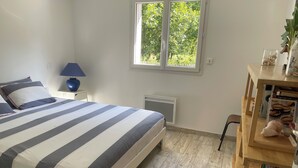 Ein Schlafzimmer mit einem Bett, einem Nachttisch, einer Lampe, einem Heizkörper, einem Fenster und einem Holzregal mit Büchern und einem Stofftier.