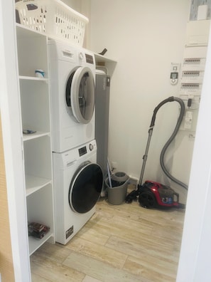 A laundry room with stacked washing machines, a vacuum cleaner, and a basket on a shelf.