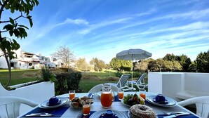 A dining table set for breakfast with croissants, fruits, and juice, overlooking a golf course and residential buildings. - ALBUFEIRA BALAIA GOLF VILAGE 1 WITH POOL by HOMING (Albufeira)