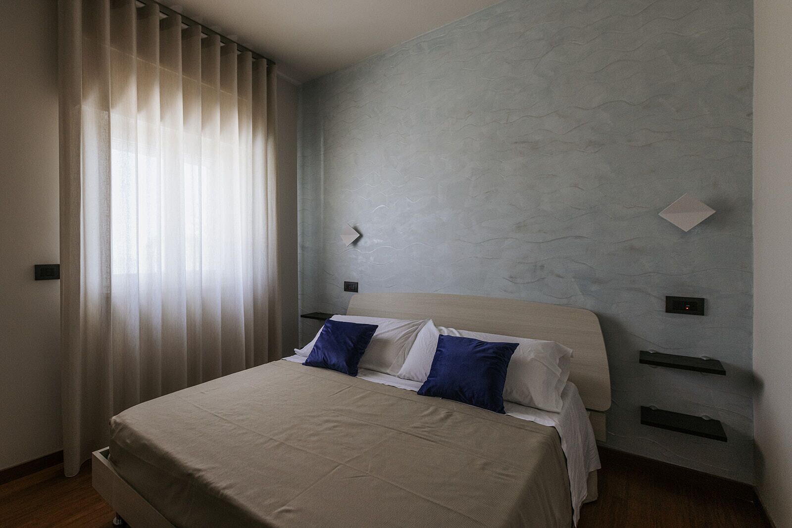 Un dormitorio moderno con cama, cortinas, pared texturizada y unidades de almacenamiento integradas.