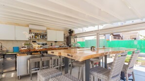An outdoor kitchen with a wooden bar, wicker chairs, and a green fence in the background. - apuliablue (Bari)