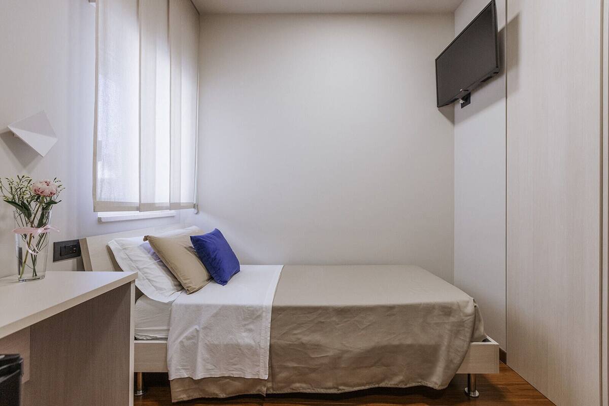 Ett litet, välordnat sovrum med en säng, ett sängbord, en tv som är monterad på väggen och ett fönster med gardiner.