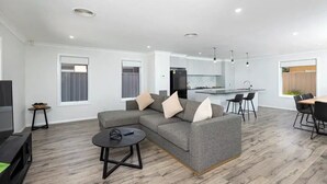 A modern living room with a grey sectional sofa, a small round coffee table, and a TV. There is a kitchen area with a fridge, a dining table with chairs, and a window with curtains. - Contemporary Living in the CBD (Wagga Wagga)