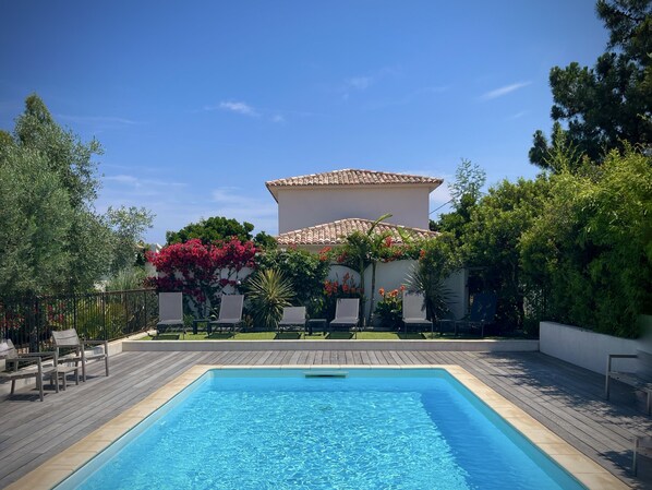 En rektangulær swimmingpool med en træterrasse hele vejen rundt, en hvid bygning med et tag af fliser i baggrunden, og frodig vegetation, herunder blomstrende planter.