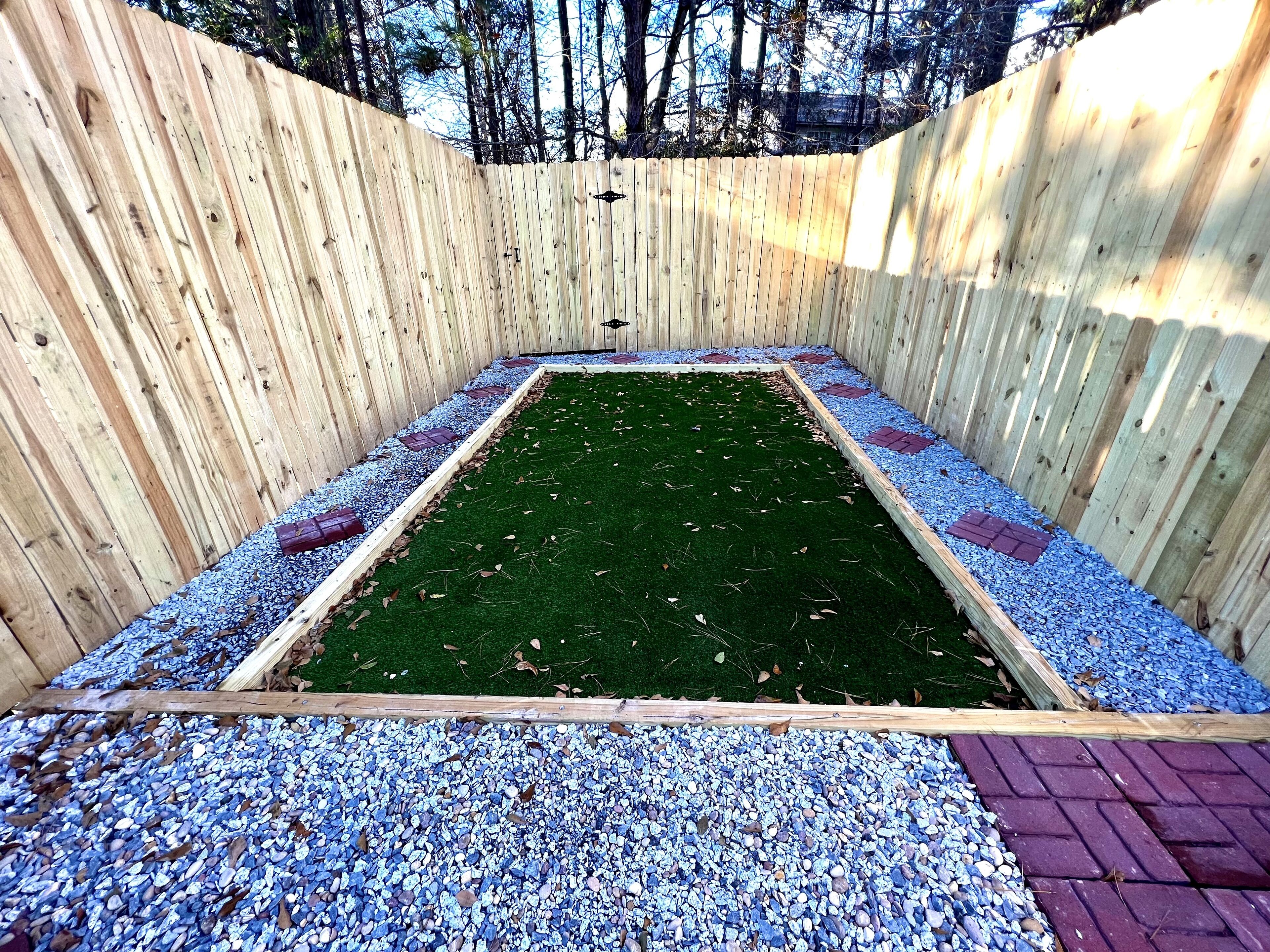 A rectangular garden area enclosed by a wooden fence with a green artificial turf and pebble ground cover.