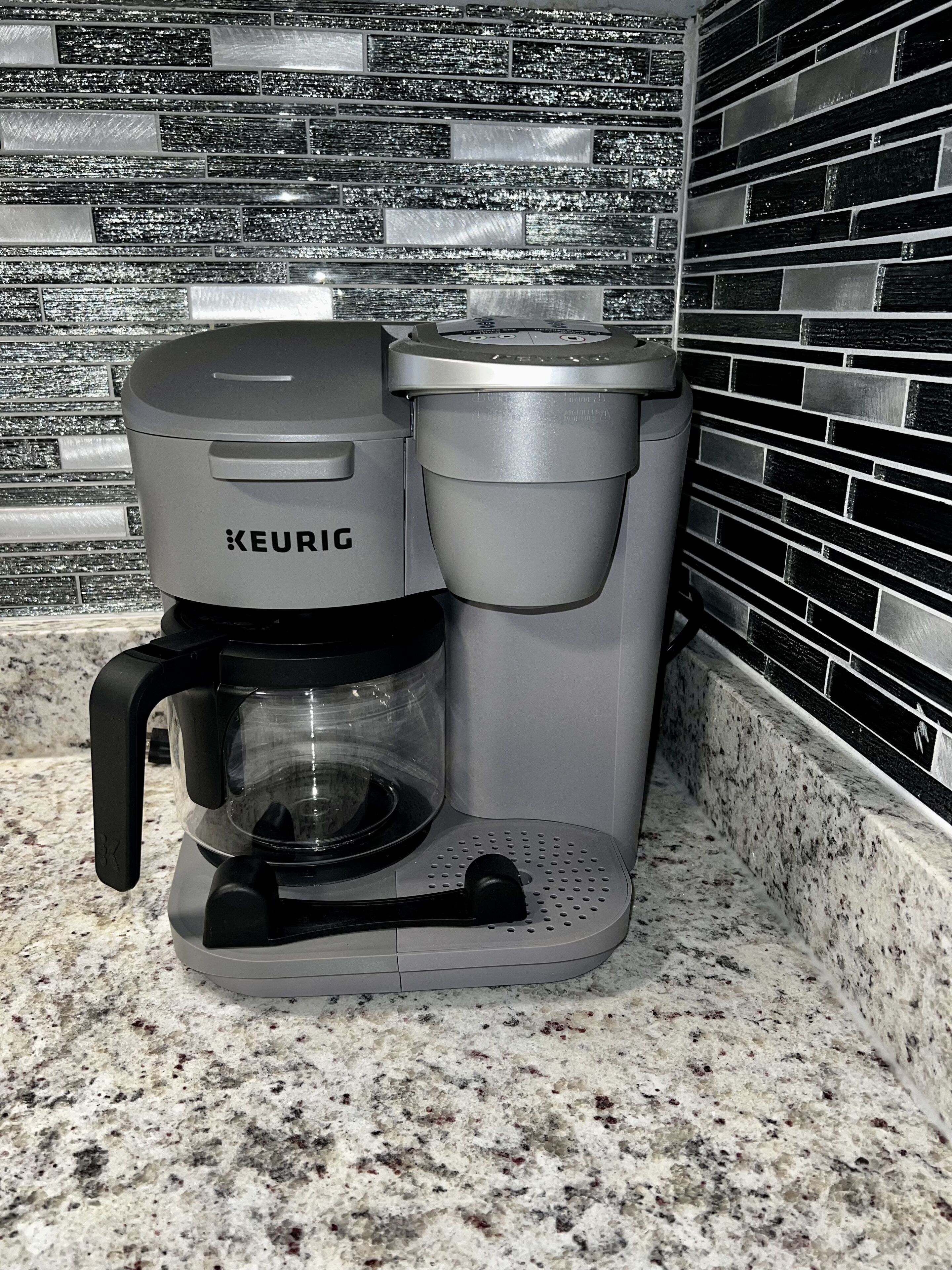 A Keurig coffee maker on a granite countertop with a black and white tiled backsplash.