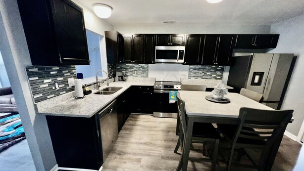 A modern kitchen with dark cabinetry, a central island, and a dining area with chairs. - 2bd 2.5 ba Sleeps 6 Luxury Townhome, Jacksonville NC. Close To Camp Lejeune (Jacksonville)