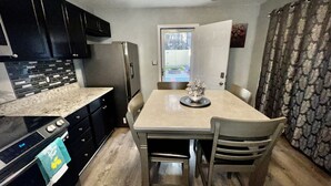 A compact kitchen with dark cabinets, a stainless steel refrigerator, a dining table with chairs, and a view through an open door. - 2bd 2.5 ba Sleeps 6 Luxury Townhome, Jacksonville NC. Close To Camp Lejeune (Jacksonville)