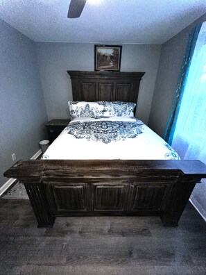 Quarto com cama de madeira escura, mesa de cabeceira, quadro na parede e janela com cortinas.