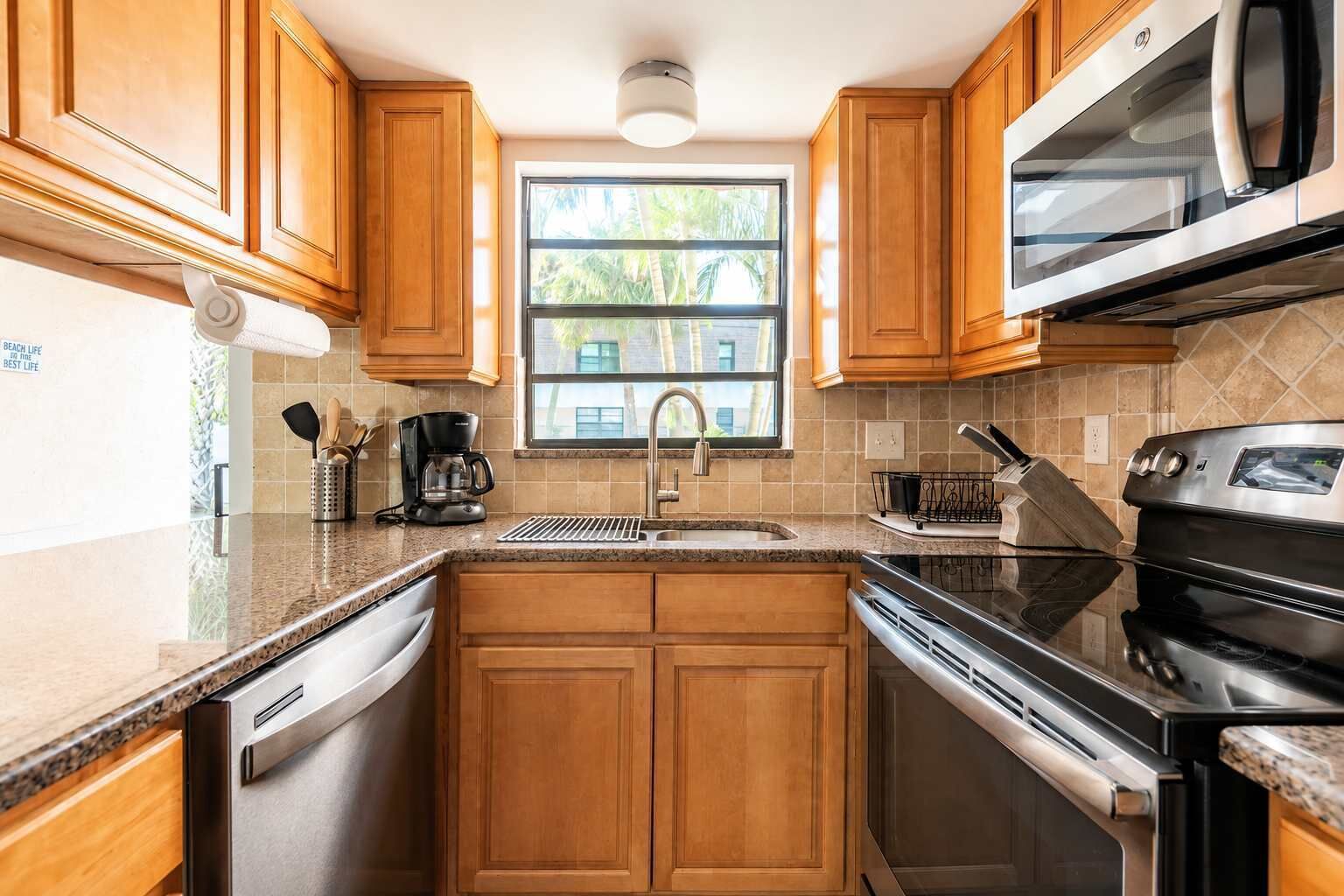 A kitchen with wooden cabinets, a stainless steel range, a microwave, and a window with a view of palm trees.
