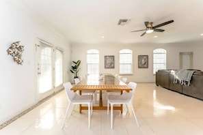 Uma sala de jantar espaçosa com uma mesa de madeira comprida e cadeiras brancas, um ventilador de teto e janelas grandes com persianas.