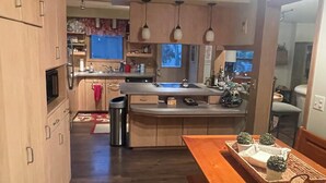 A modern kitchen with wooden cabinets, a central island, pendant lights, and a dining area with a wooden table. - Beautiful waterfront home on Mickey Lake, channeling to 2800 acre Long Lake. AC  (Traverse City)