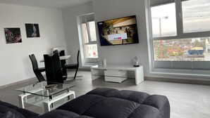 Uma sala de estar moderna com uma mesa de centro de vidro, um sofá, uma TV fixada na parede e vista para uma obra em construção pela janela.