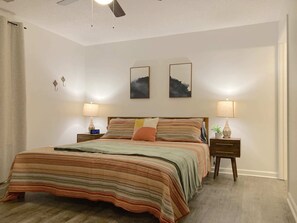Un dormitorio con una cama, dos mesitas de noche, un tocador, un ventilador de techo y dos obras de arte colgadas en la pared.
