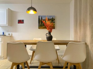 Uma área de jantar com uma mesa de madeira, cadeiras brancas, um vaso preto com flores, um quadro na parede e um aviso de 'Não Fumar'.