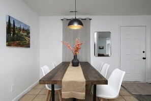A dining area with a long table and comfortable seating, featuring a pendant light, a vase with flowers, and a beautiful landscape painting.