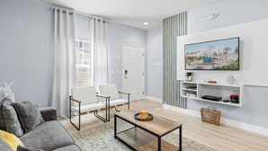 Un salon moderne avec un canapé gris, des fauteuils blancs, une table basse en bois et un téléviseur fixé au mur qui diffuse une scène des studios Universal.