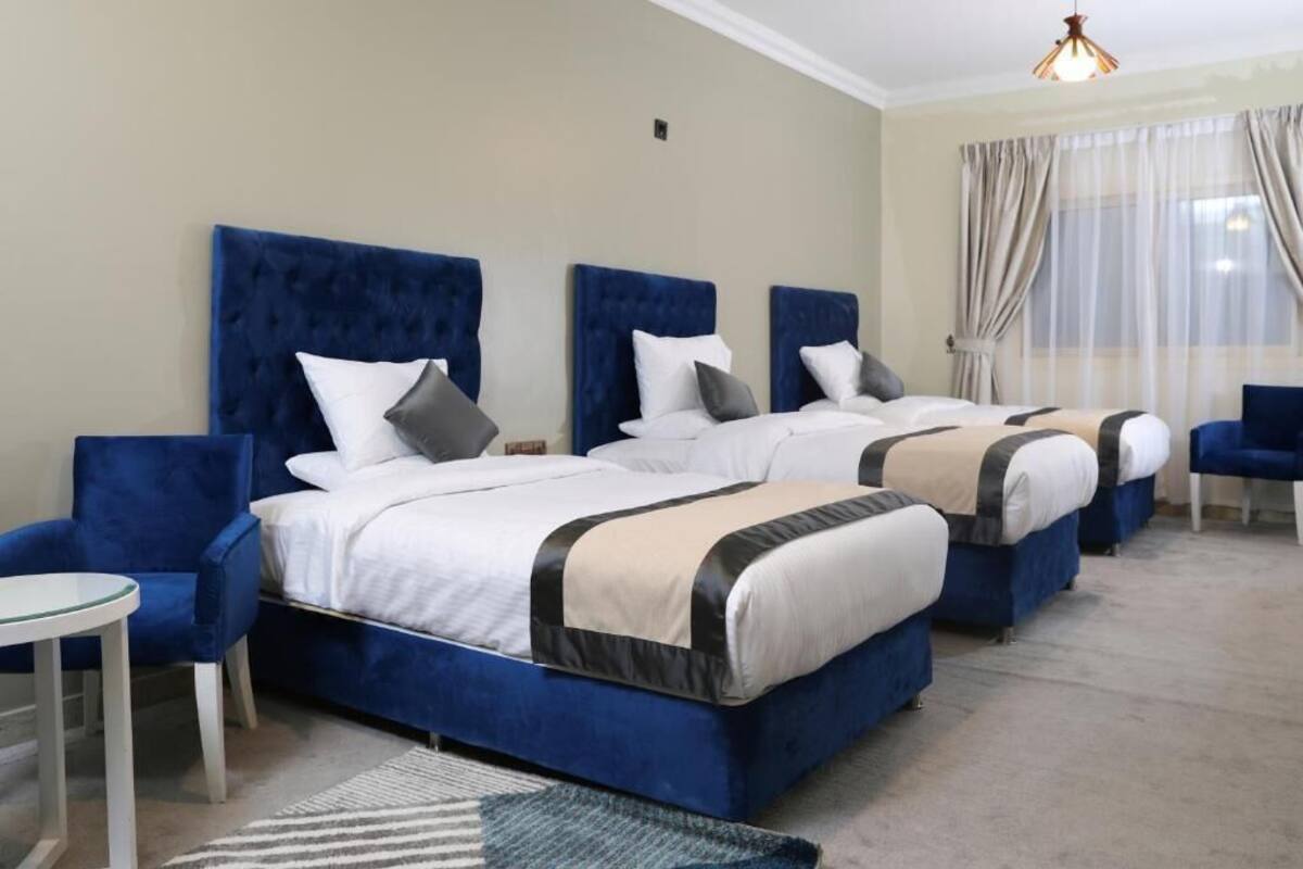 Habitación de hotel con dos camas, cada una con cabecera azul, ropa de cama blanca y alfombra a rayas. Hay una mesita redonda y una silla azul en la habitación.