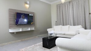 Una sala de estar moderna con un televisor de pantalla plana montado en la pared, una mesa de centro de vidrio, sofás blancos y un ventanal con cortinas.