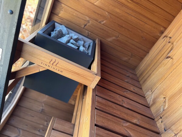 Un banco de madera con una caja negra que contiene rocas, etiquetada como "FINNTHERM", en un ambiente de cabaña de madera.