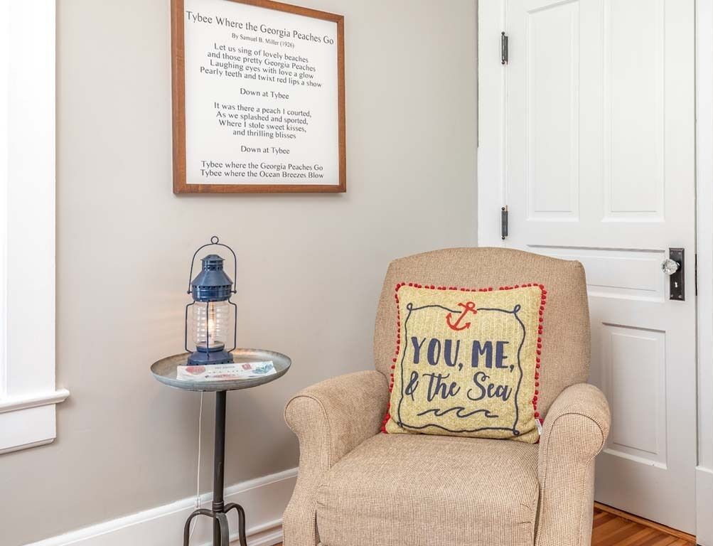 A room with a framed poem on the wall, a small table with a lantern, and a chair with a cushion that says 'YOU, ME, & the Sea'.