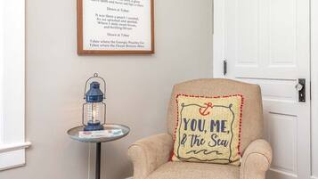 A room with a framed poem on the wall, a small table with a lantern, and a chair with a cushion that says 'YOU, ME, & the Sea'.