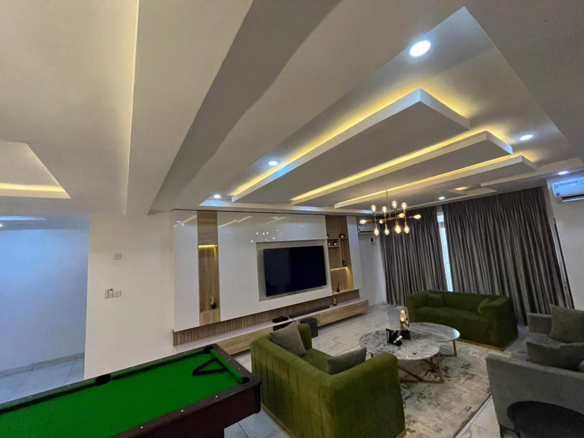 A modern living room with a pool table, green sofas, a coffee table, and a flat-screen TV.