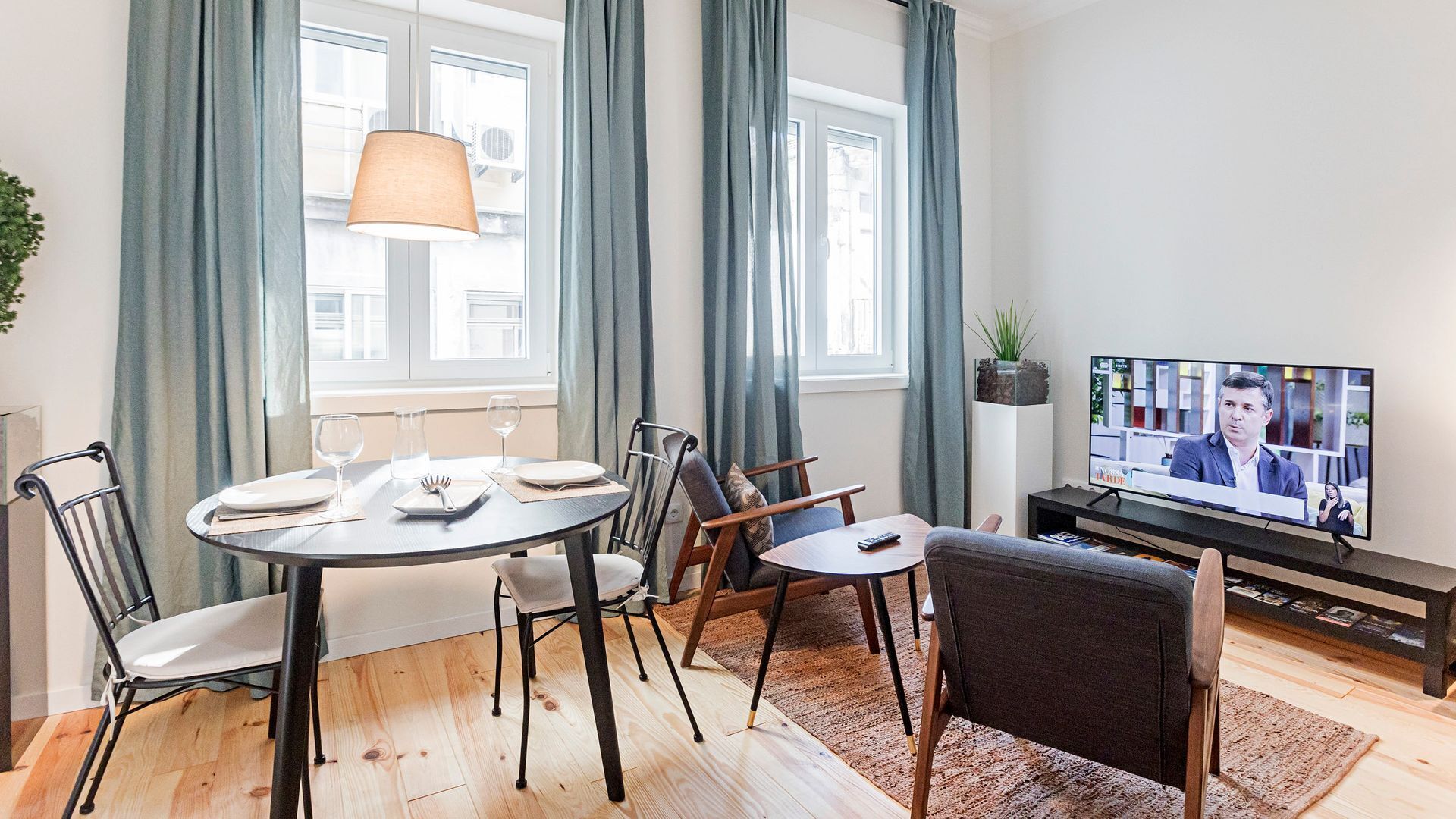 Ein modernes Wohnzimmer mit Esstisch für zwei Personen, einer Couch und einem Fernseher, der eine Nachrichtensendung zeigt.