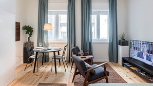 Een moderne woonkamer met een eethoek, grote ramen voorzien van gordijnen, een televisie en een boekenkast.