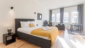 A modern bedroom with a large bed, a bedside table, a small desk, a chair, and a view of the outdoors through large windows. - 150 Rivoli Apartments - 3ºA by LovelyStay (Porto)