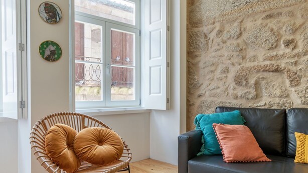A room with a wicker chair, a black leather sofa with colorful cushions, a stone wall, and a window with a view of a balcony. - Riverside Cozy Studio by LovelyStay (Porto)