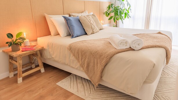 Een net opgemaakt bed met een beige deken en kussens, een klein houten nachtkastje met een plant en boeken, en een groot raam met natuurlijk licht.