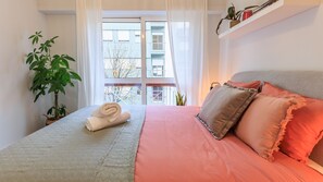 A neatly made bed with a rolled towel, a potted plant, and a view of a building through a window.