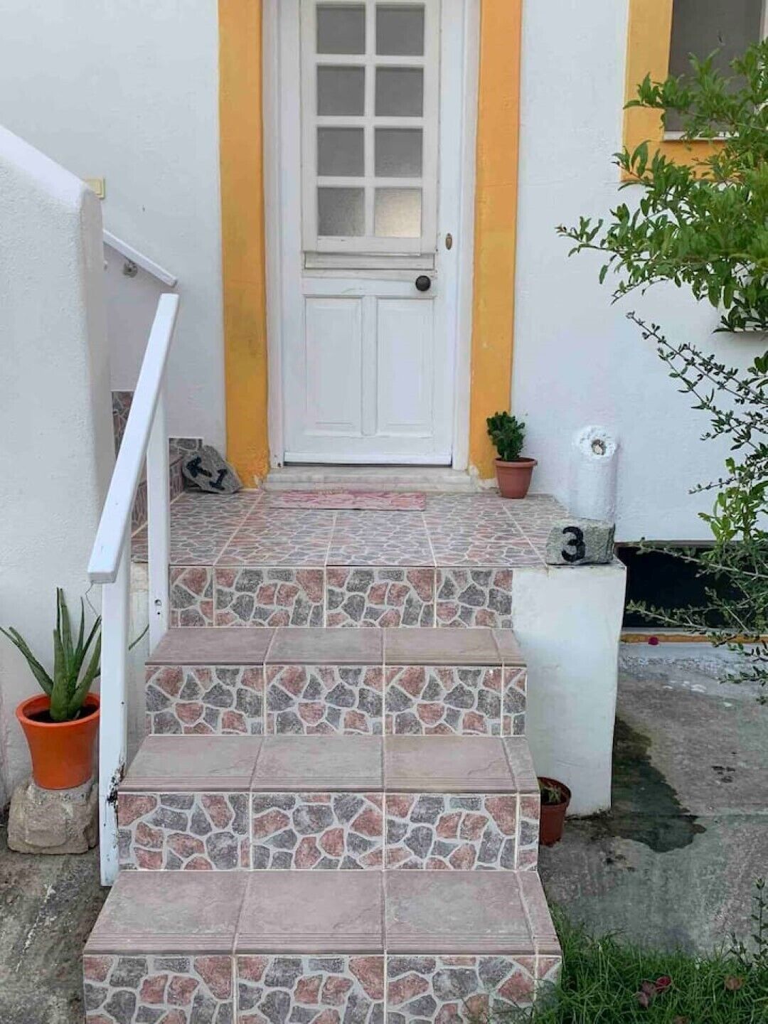 Un conjunto de escalones de piedra con baldosas que conducen a una puerta blanca con un panel de vidrio, flanqueada por una pared amarilla y una planta en maceta.