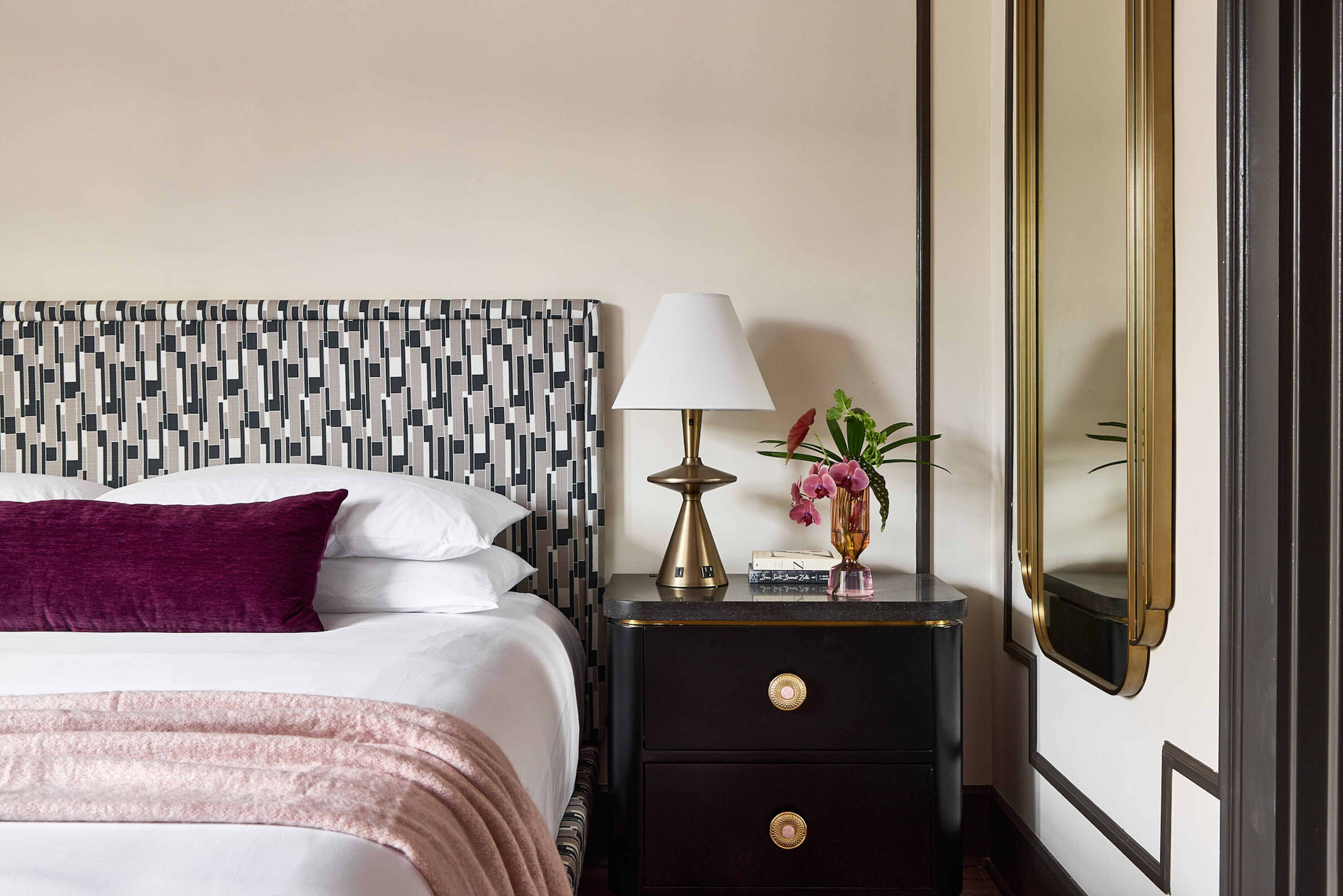 A neatly made bed with white linens and a pink throw blanket, a black nightstand with a gold lamp, and a mirror on the wall.